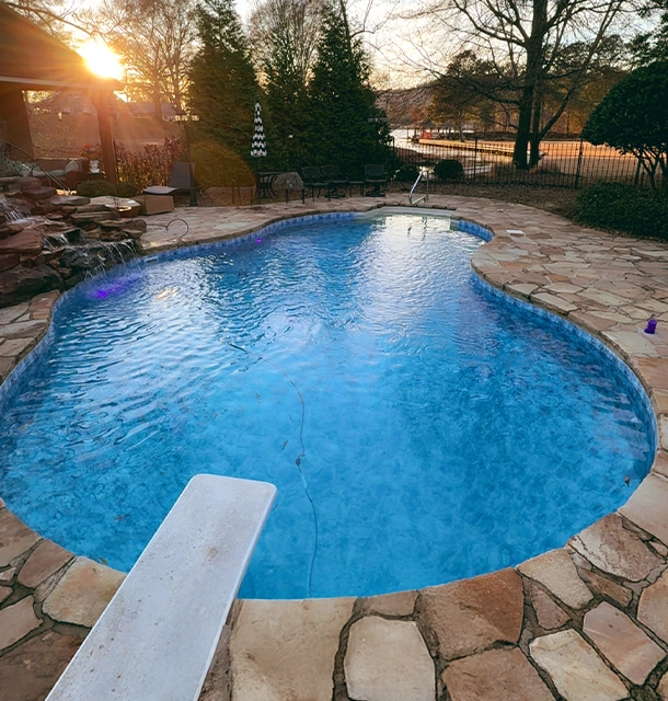 kidney-shaped pool with diving board surrounded by stone patio at sunset